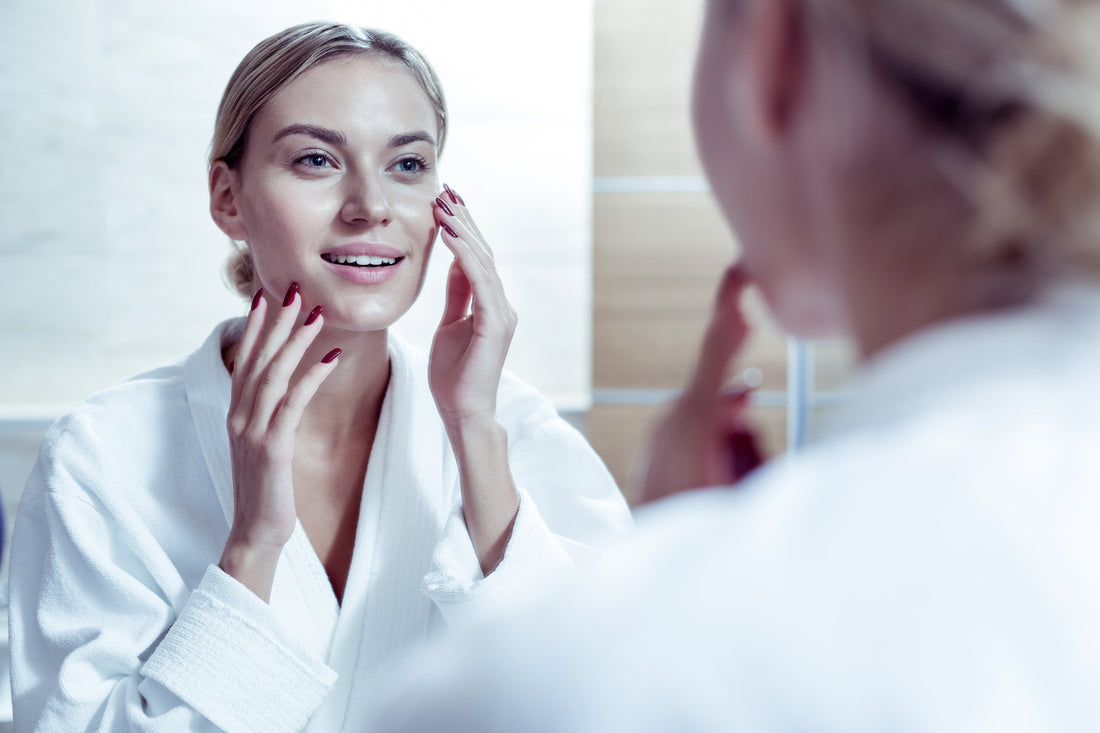 Femeie în halat alb își examinează tenul în oglindă, moment din rutina zilnică de îngrijire a pielii.