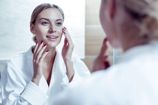 Femeie în halat alb își examinează tenul în oglindă, moment din rutina zilnică de îngrijire a pielii.