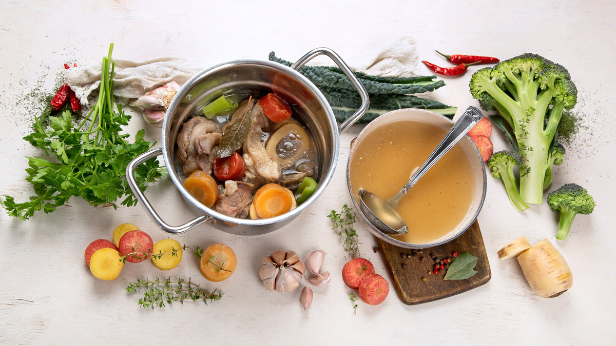 Oală cu supă din oase și legume, alături bol cu supă clară; ingrediente proaspete (broccoli, usturoi, ierburi) pe masă — scenă de bucătărie pentru supă de oase.
